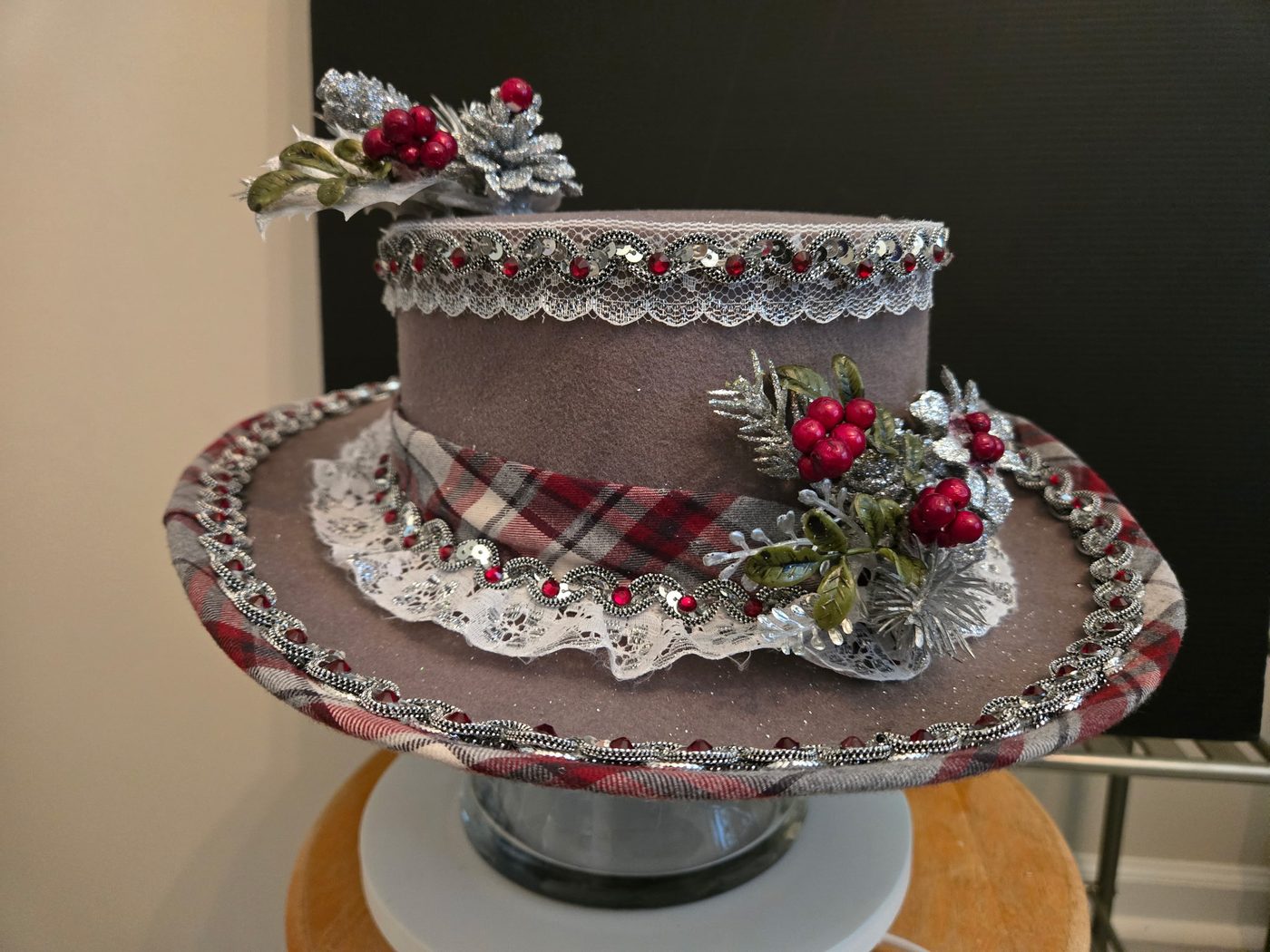 Plaid Hatters tartan top hat trimmed with lace, sequins, holly berries, and silvered pine sprigs, from the 2026 Christmas Collection.